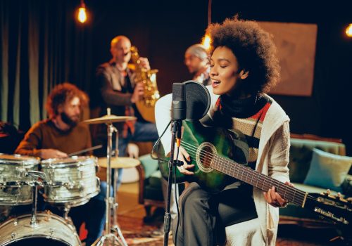 Mixed race woman singing and playing guitar while sitting on chair with legs crossed. In background drummer, saxophonist and bass guitarist.